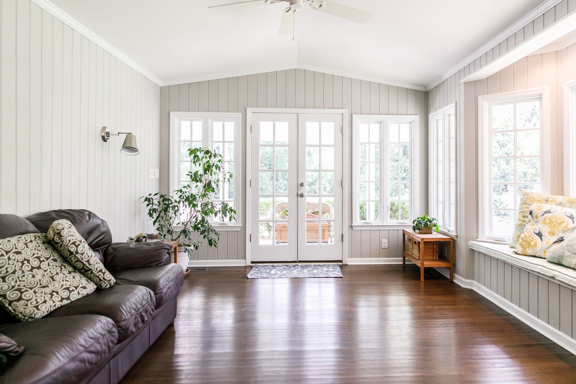 Bright sunroom with white walls, dark sofa, potted plants, French doors, and hardwood floors