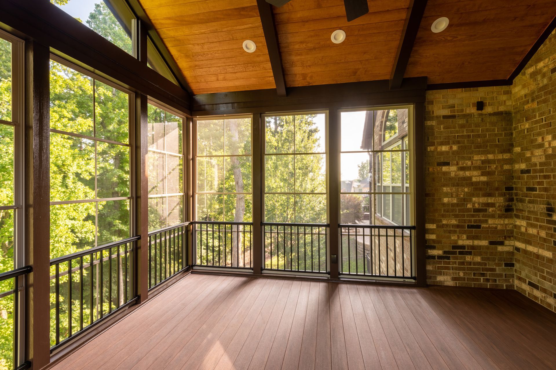 Sunroom with wood-paneled ceiling, brick wall, and large windows overlooking green trees