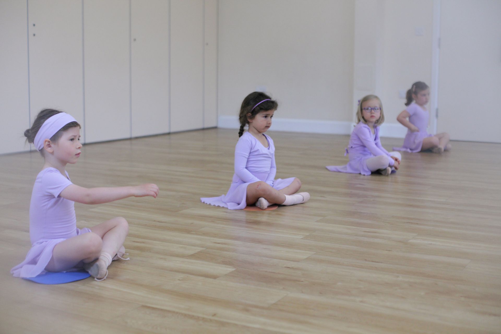 Four young children in lilac dance outfits sit on a wooden floor, arms extended, in a dance studio.