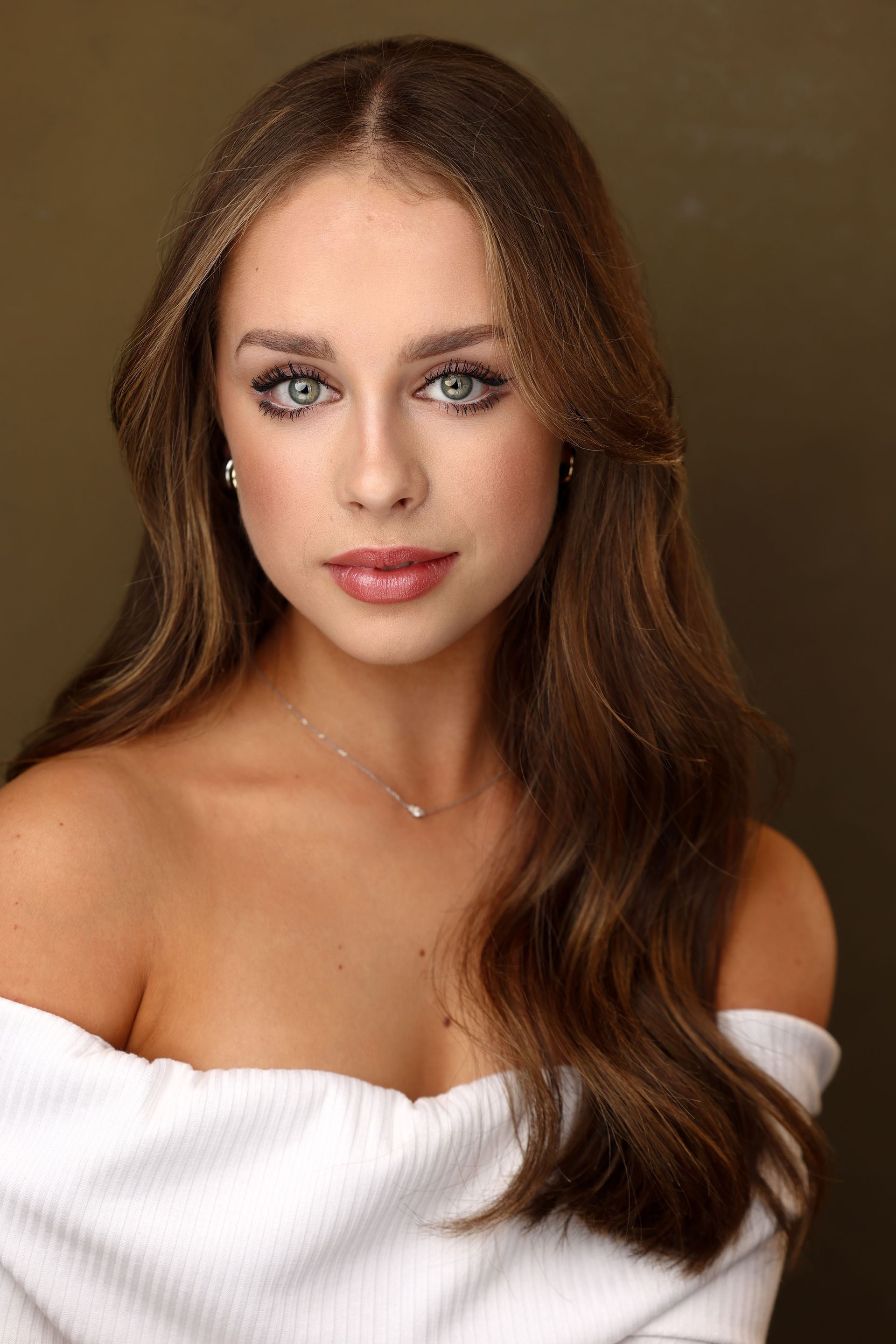 Woman with long wavy brown hair and blue eyes, wearing an off-the-shoulder white top, looking at the camera.
