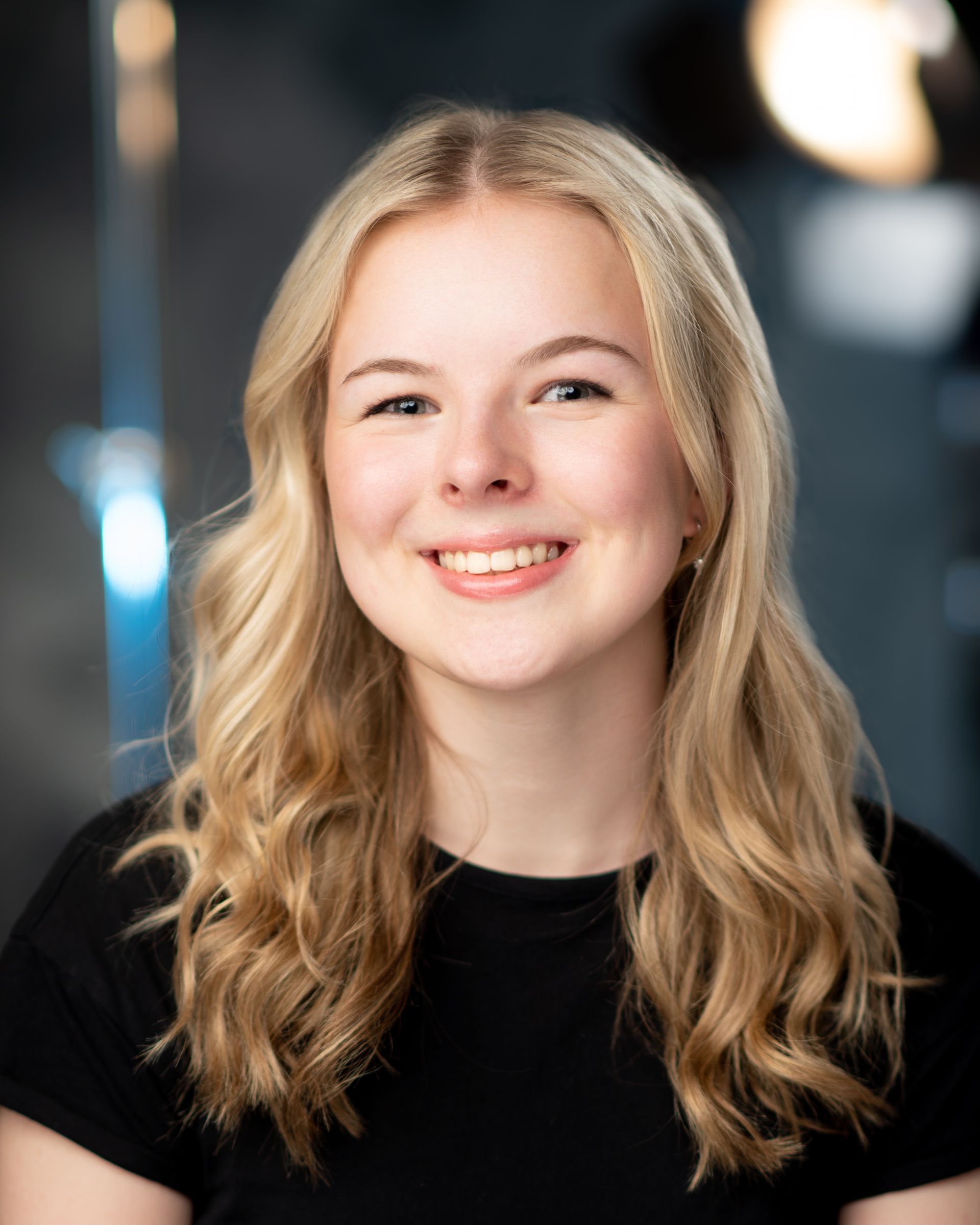 Blonde woman with a bright smile, wearing a black shirt, posing in front of a blurred background.