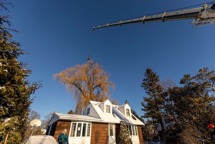 A house with a snowy roof, trees, and a crane against a bright blue sky. A person stands near the house.