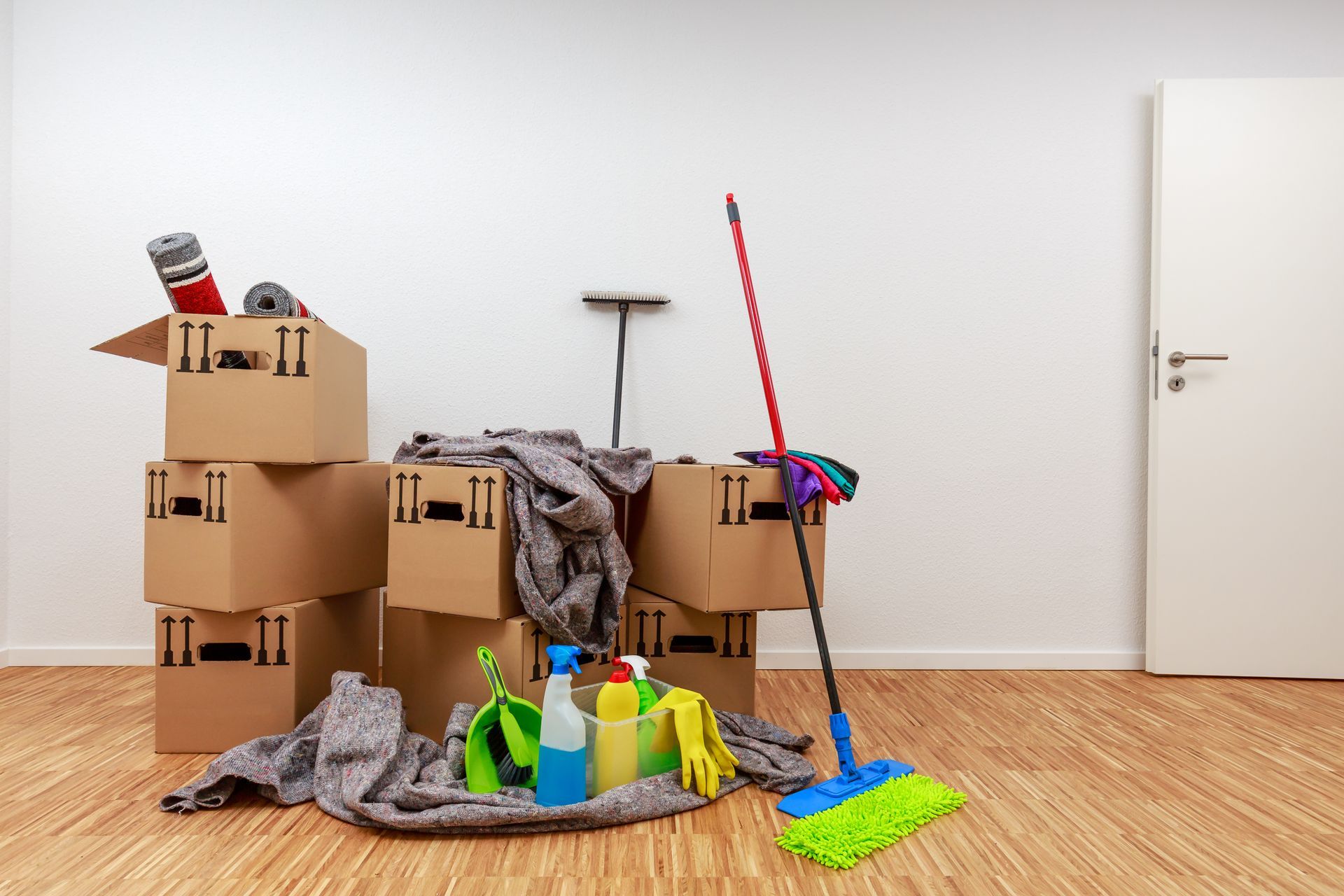 Cardboard boxes, cleaning supplies, and mop in room with wooden floor and white door.