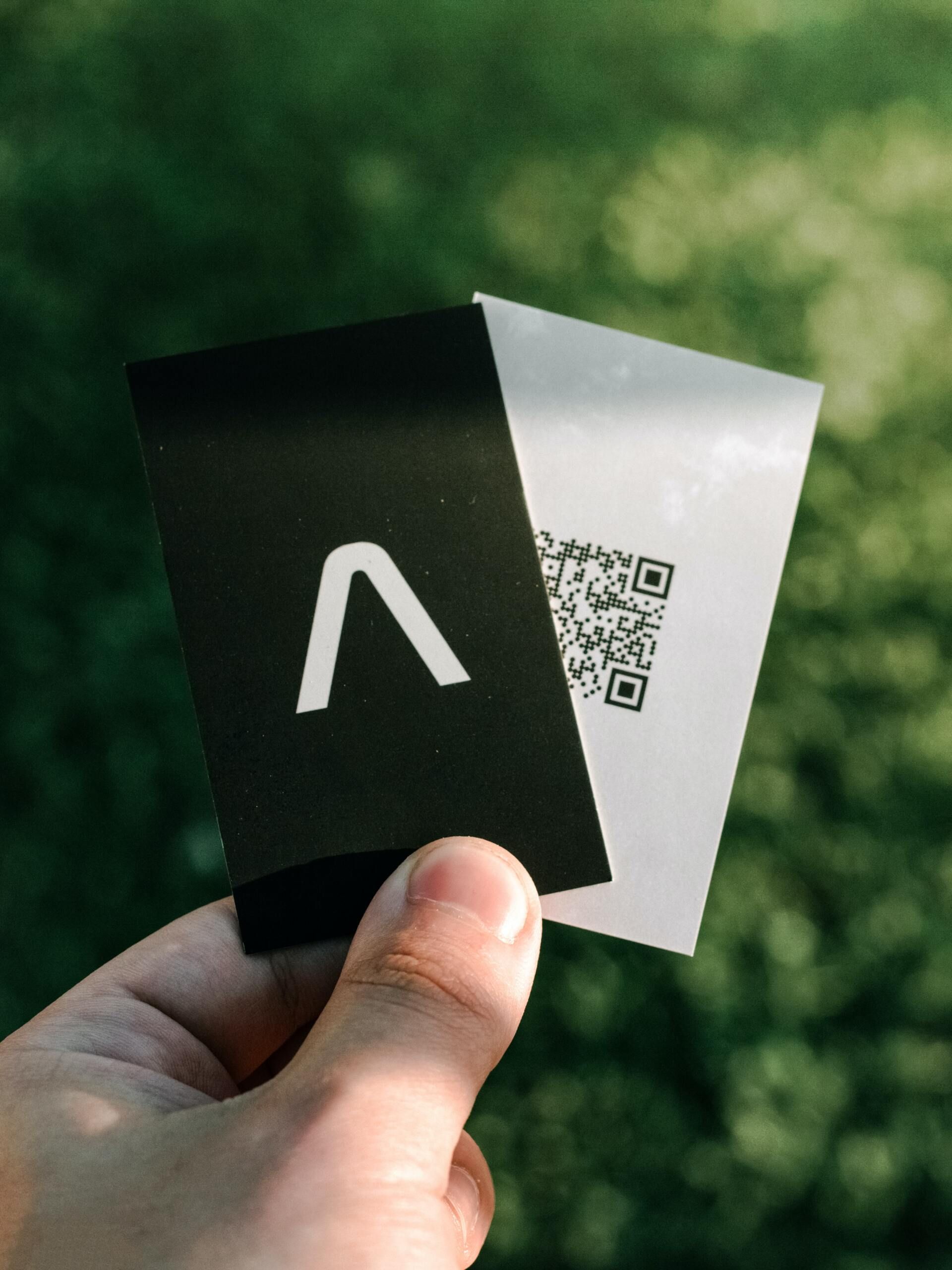 Una mano sostiene dos tarjetas de visita sobre un fondo verde suave: una negra con un logotipo blanco y otra blanca con un código QR.