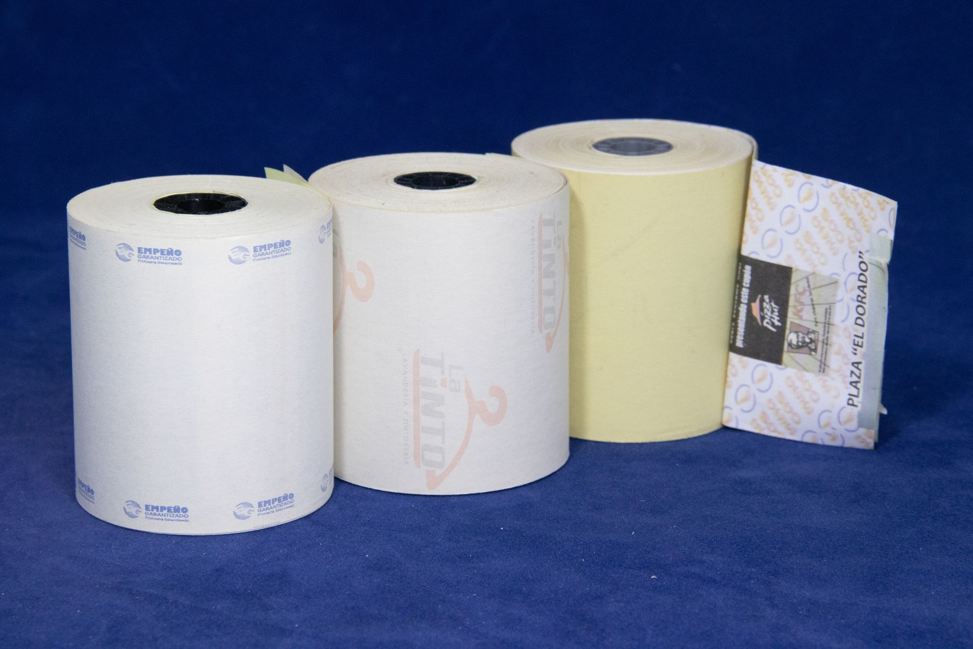 Tres rollos de papel, blanco, blanquecino y amarillo, sobre una superficie azul. Junto a ellos hay un recibo doblado.