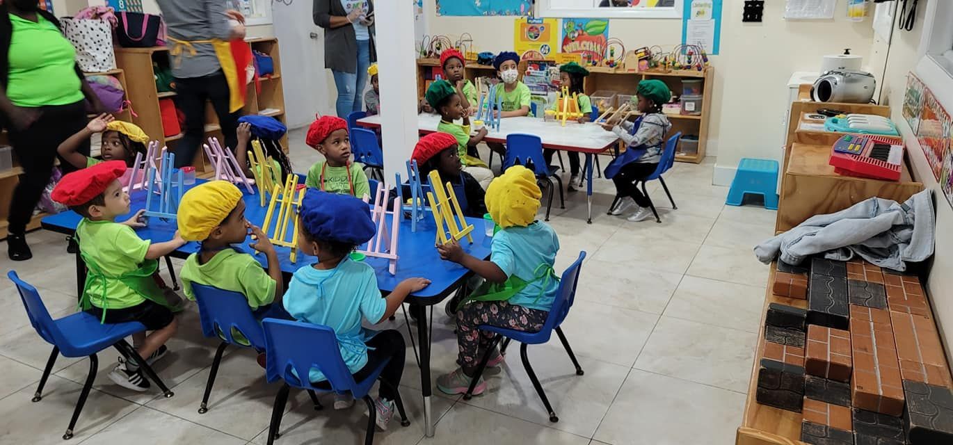 Children in colorful berets sit at blue tables in a classroom, crafting with tall, yellow structure pieces.