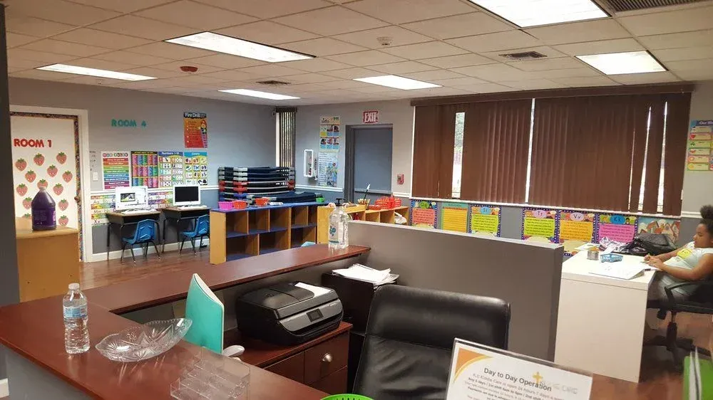 An office reception area with a desk, printer, chairs, and cubicle-style partitions, with an open room in the background.