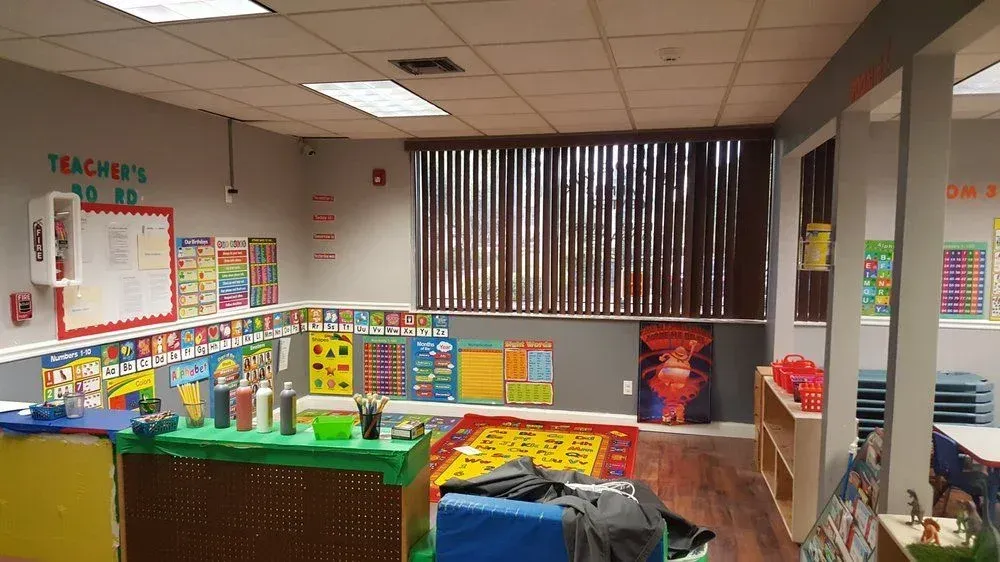 A colorful, organized preschool classroom with learning posters, a green activity table, and a rug on a hardwood floor.