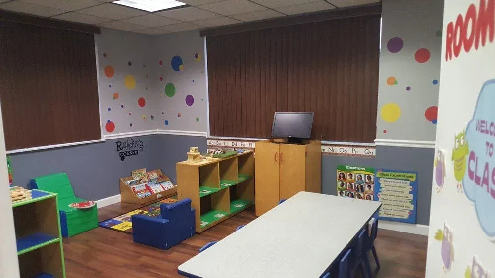 A colorful, tidy classroom with a long table, chairs, storage shelves, books, and wall decorations on gray walls.