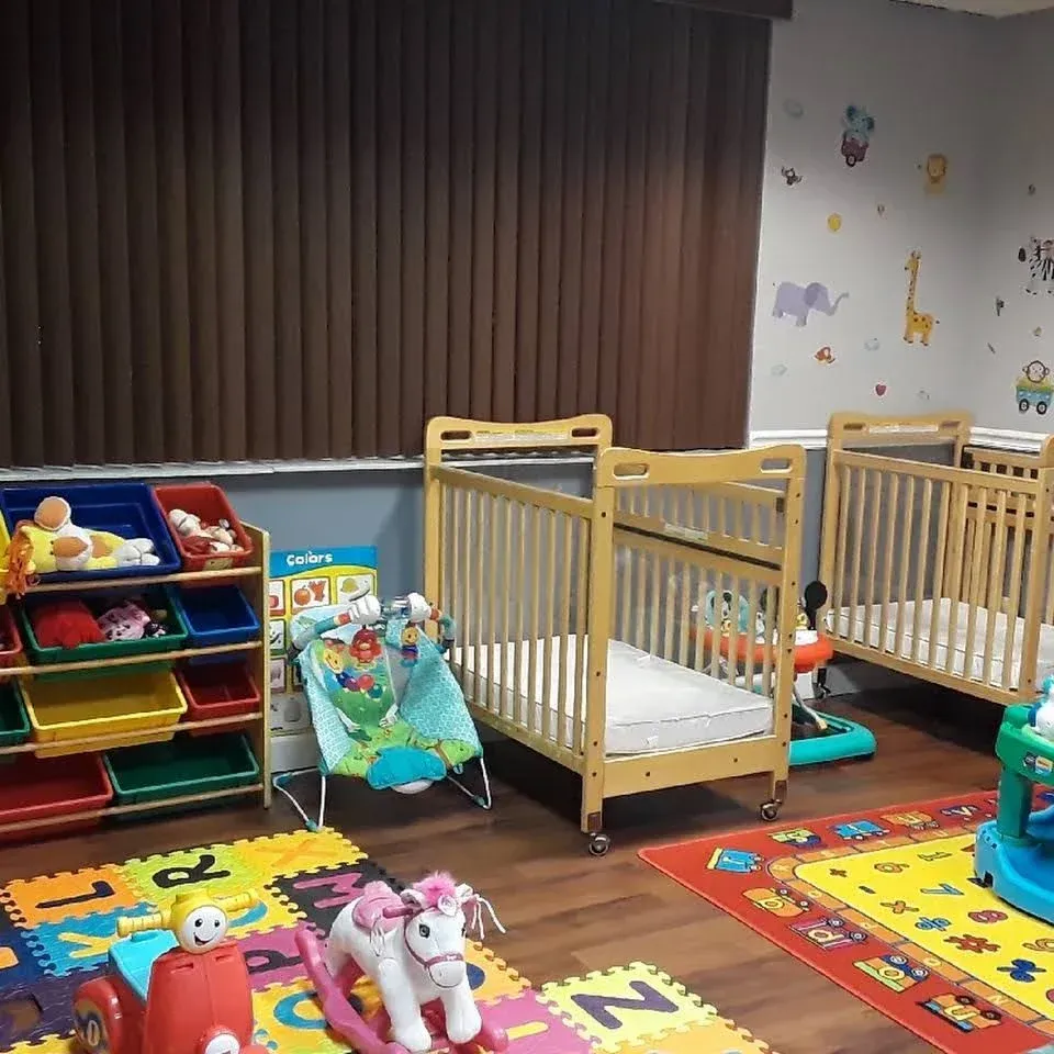 A bright childcare room features wooden cribs, toy storage bins, play mats, and various toys for children.