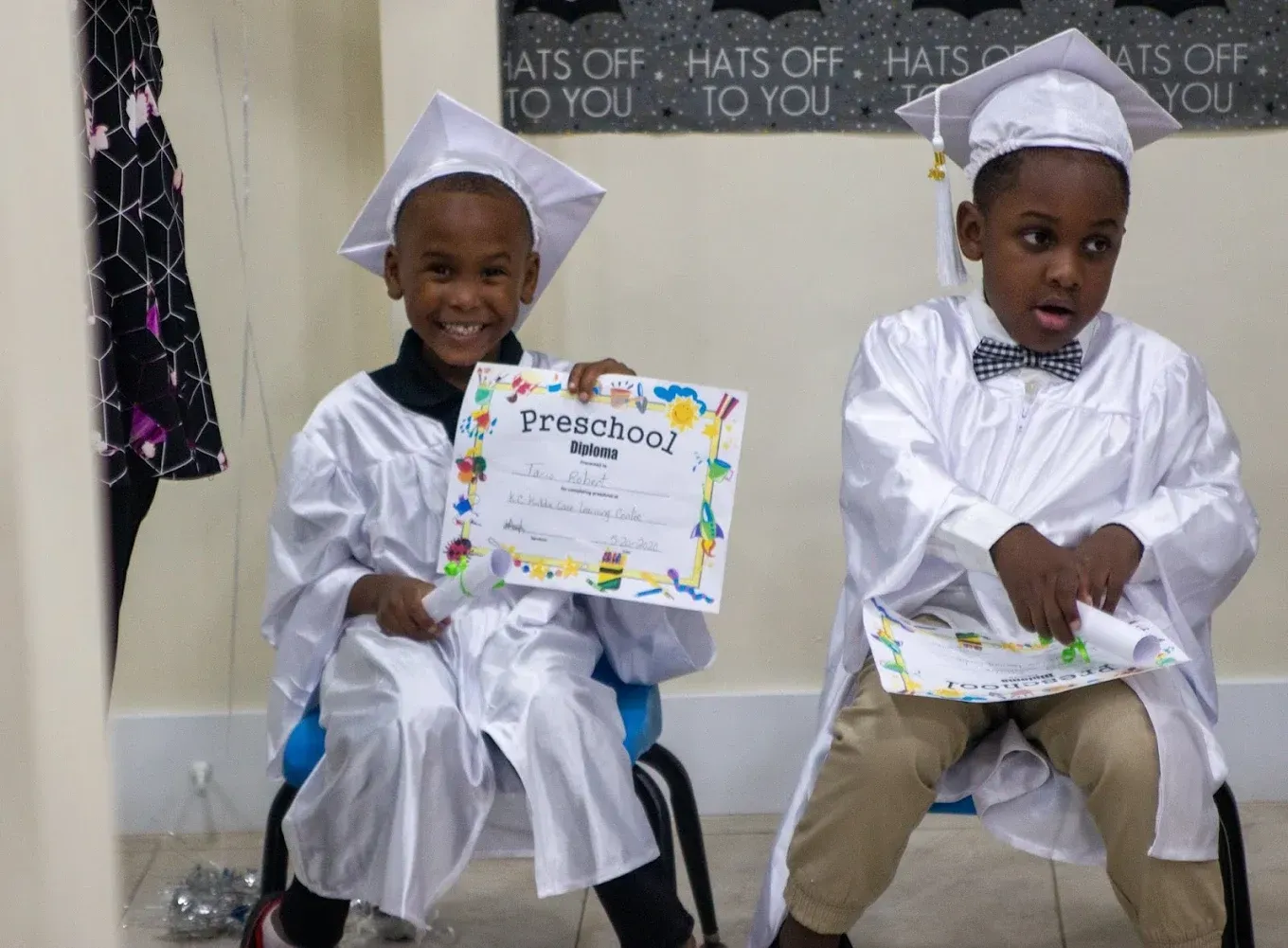 Two children in white graduation gowns and caps sit on chairs, one smiling and holding a preschool diploma.