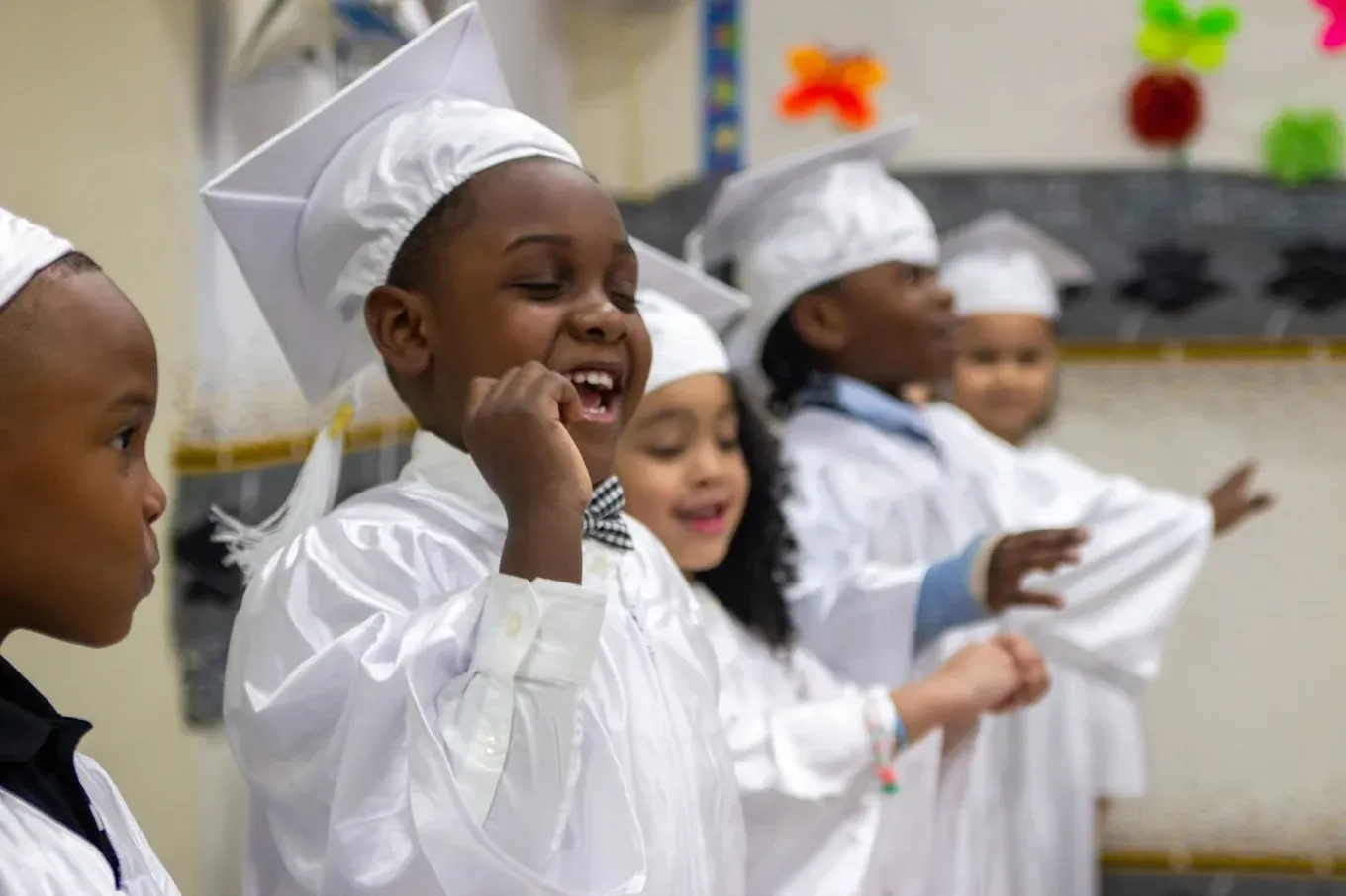 Children in white graduation caps and gowns standing together, smiling and performing on stage.