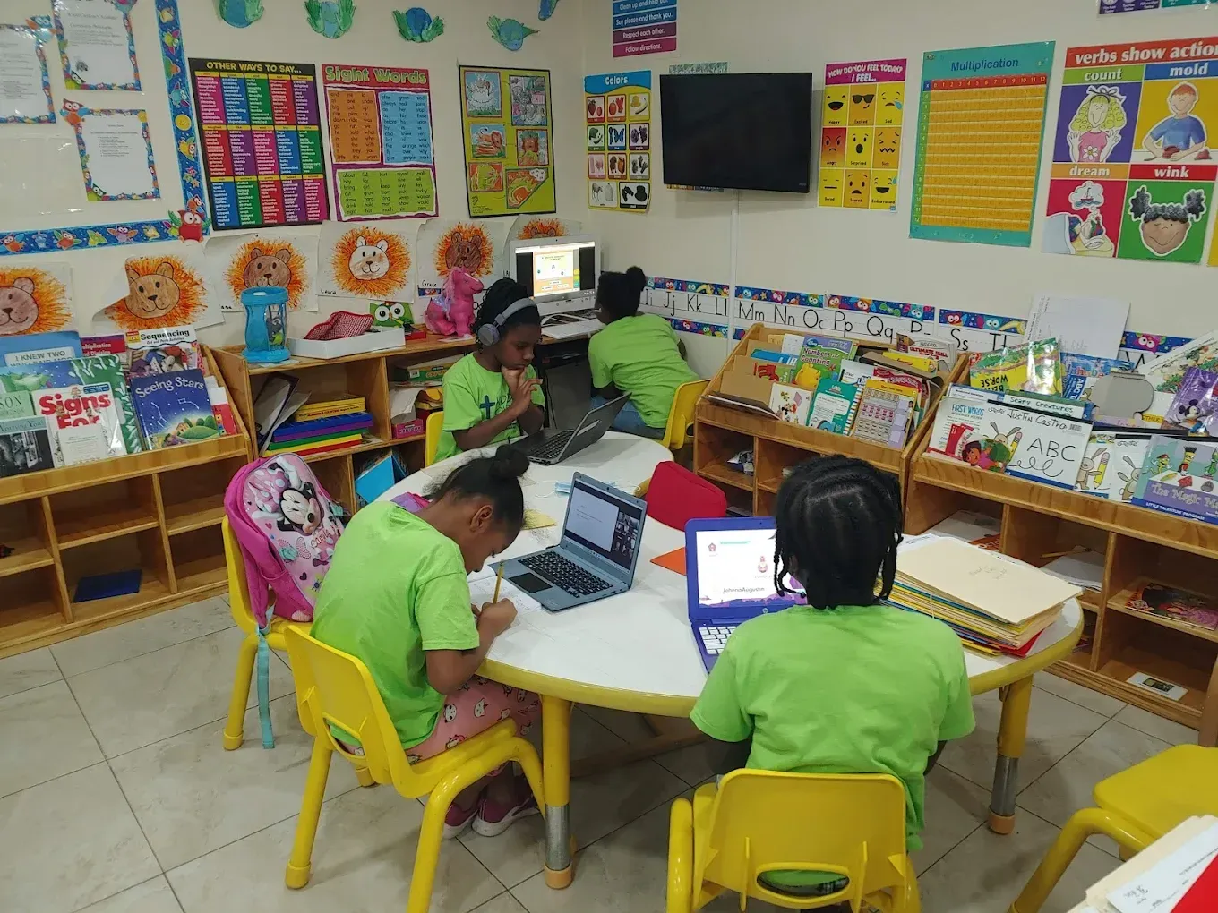 Children in lime green shirts use laptops at a round table in a classroom filled with educational posters and bookshelves.
