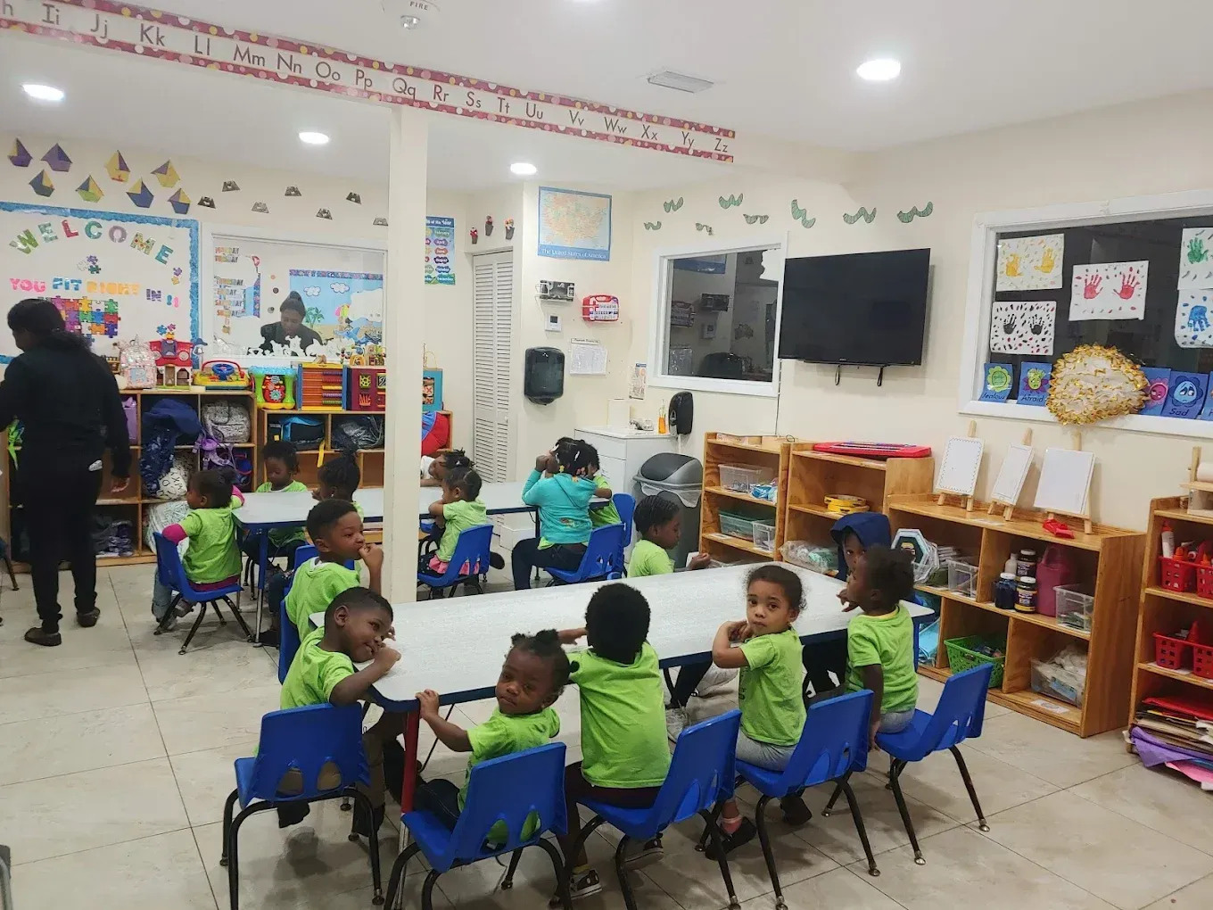 A brightly lit classroom with children in green shirts sitting at tables, with an adult standing to the left.