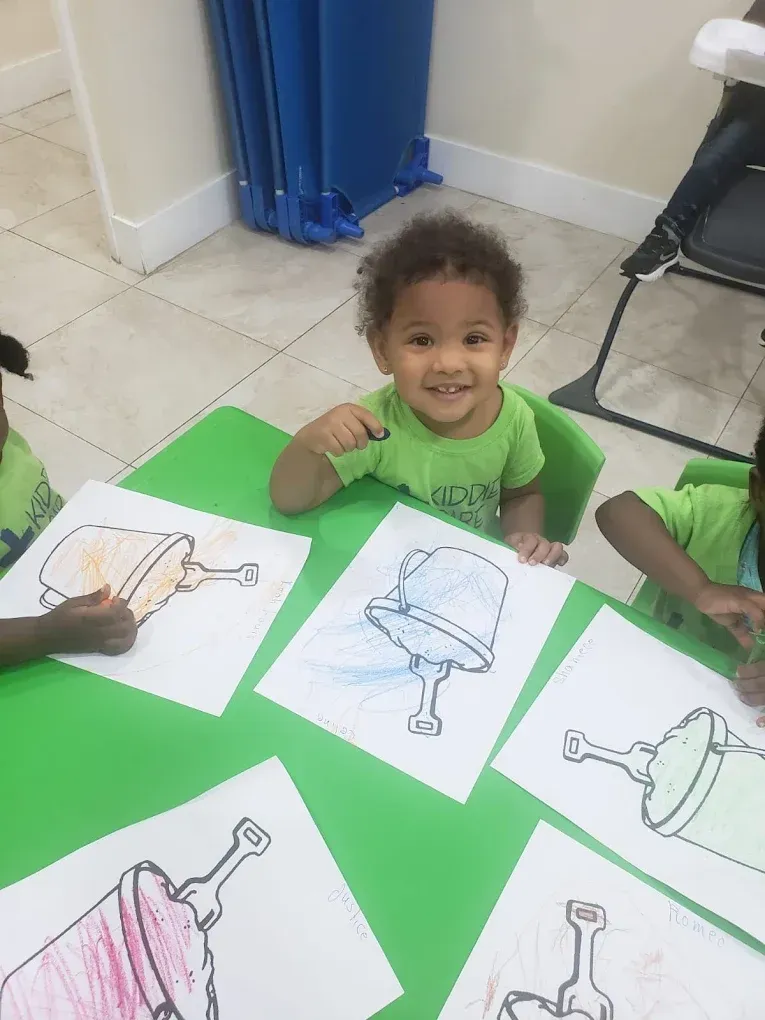 A smiling child sits at a green table, coloring pictures of sand buckets and shovels in a classroom setting.