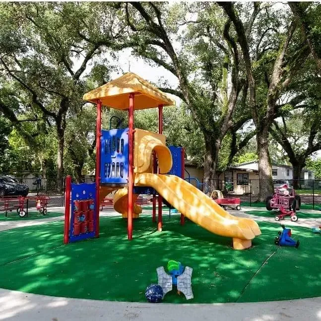A yellow and blue playground structure with a slide on artificial green turf under large trees, with toys scattered nearby.