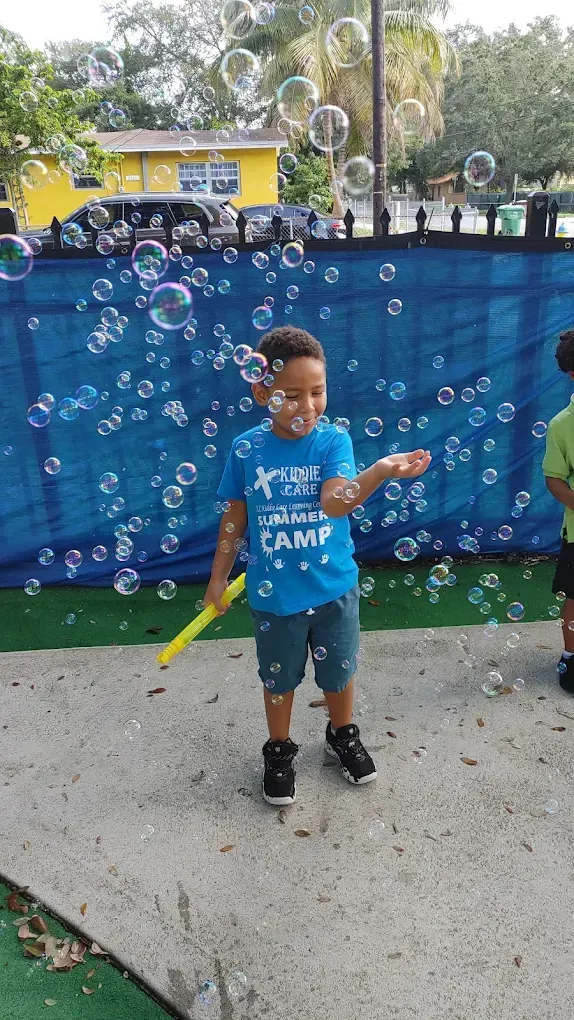 A smiling child in a blue t-shirt plays with bubbles in an outdoor area with a blue fence in the background.
