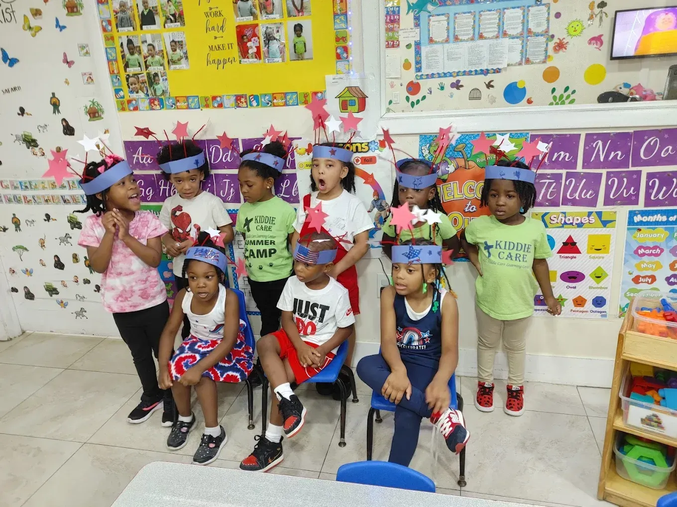 A group of children wearing homemade star-decorated headbands stand in a classroom decorated with educational posters.