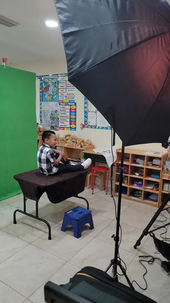 A person sits on a table in front of a green screen in a classroom, with photography lighting equipment nearby.