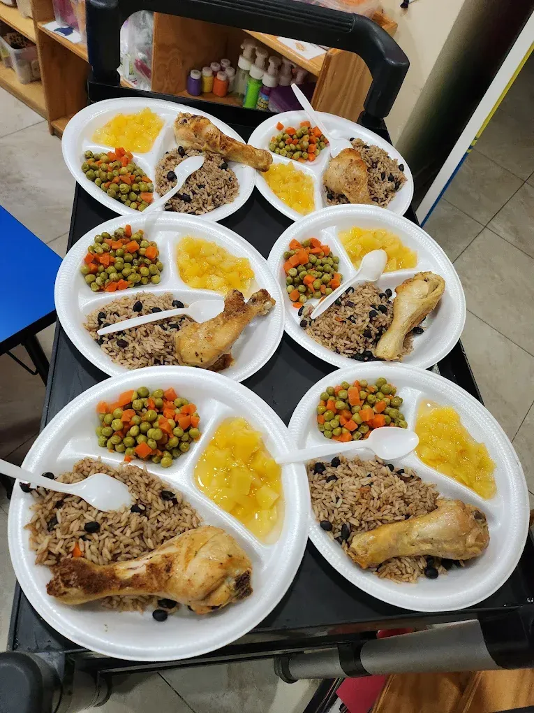 Six white divided plates on a cart, each containing a chicken leg, rice and beans, mixed vegetables, and yellow fruit.