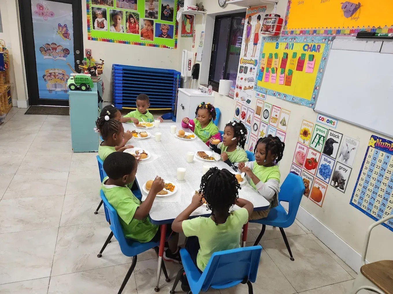 Children in green shirts sit around a classroom table eating lunch on blue chairs in a brightly decorated room.