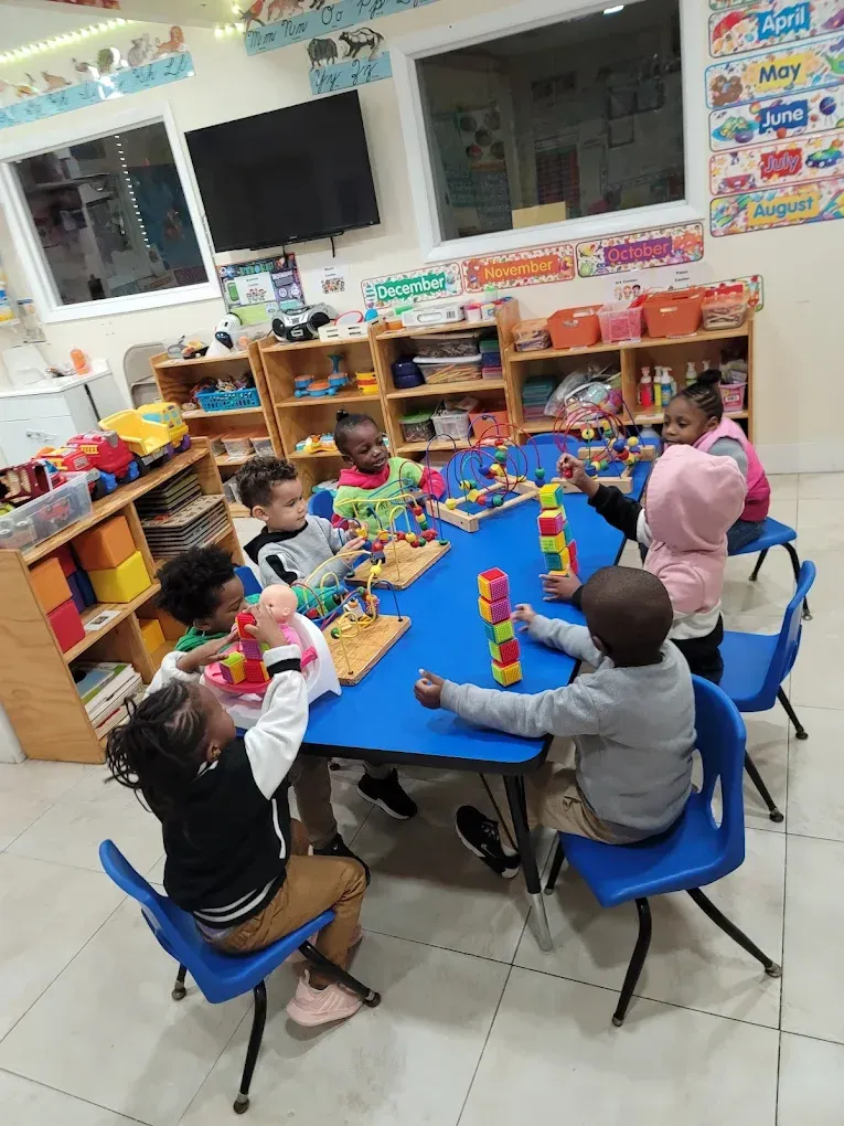 Children sit at a blue table in a colorful classroom, building with blocks and playing with toys.