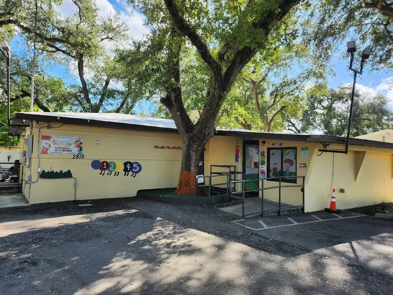A yellow one-story building with a large tree in front, a metal ramp at the entrance, and parking spots in the foreground.