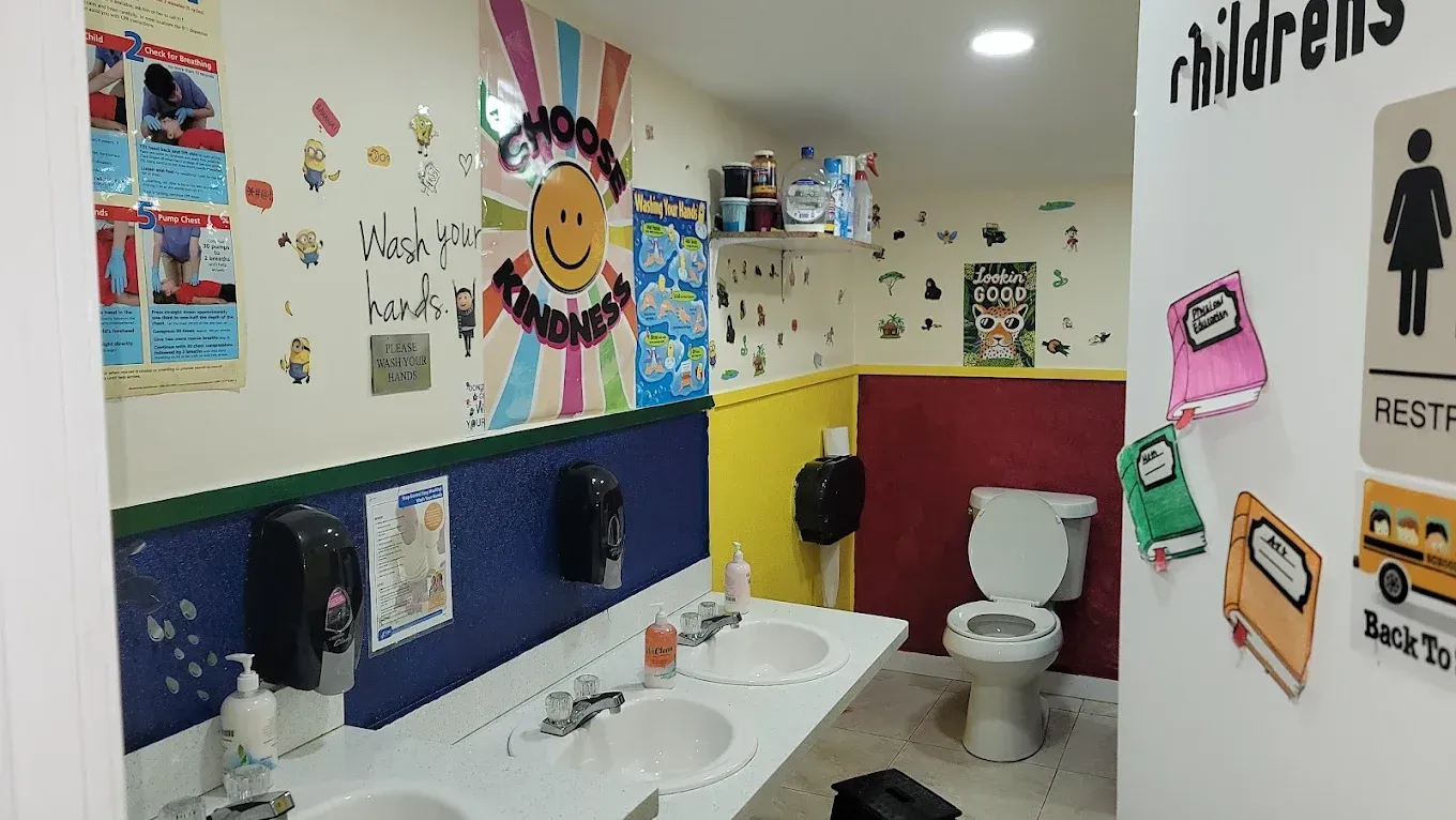 A colorful children's bathroom featuring two sinks, a toilet, and walls decorated with posters and educational stickers.