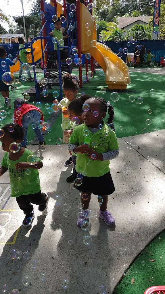 Children playing with bubbles on a sunny playground near a yellow slide.
