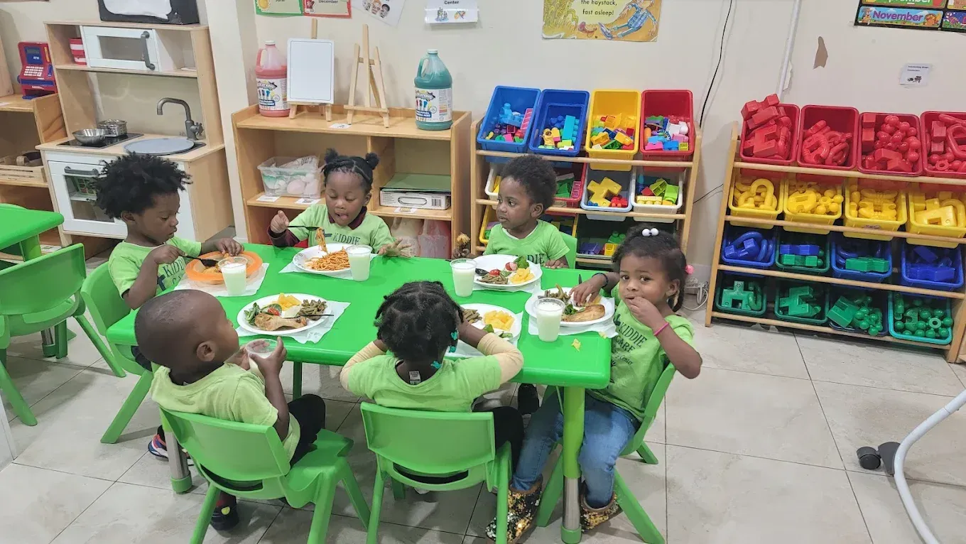 Children in matching green shirts sit around a table in a bright classroom eating lunch and drinking milk.