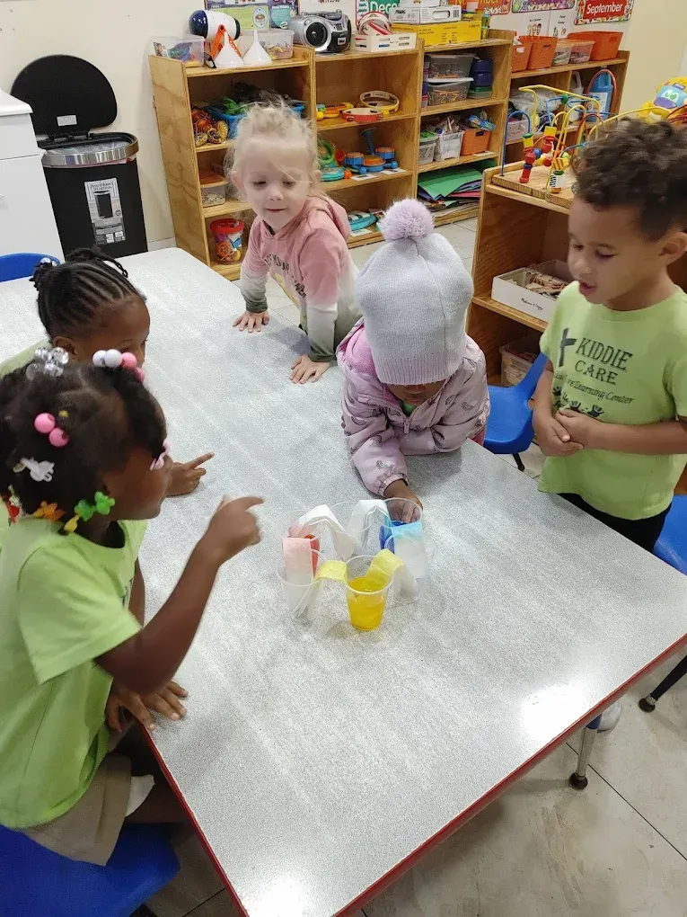 Five children gather around a table in a classroom, pointing and looking at several plastic cups filled with colored liquid.