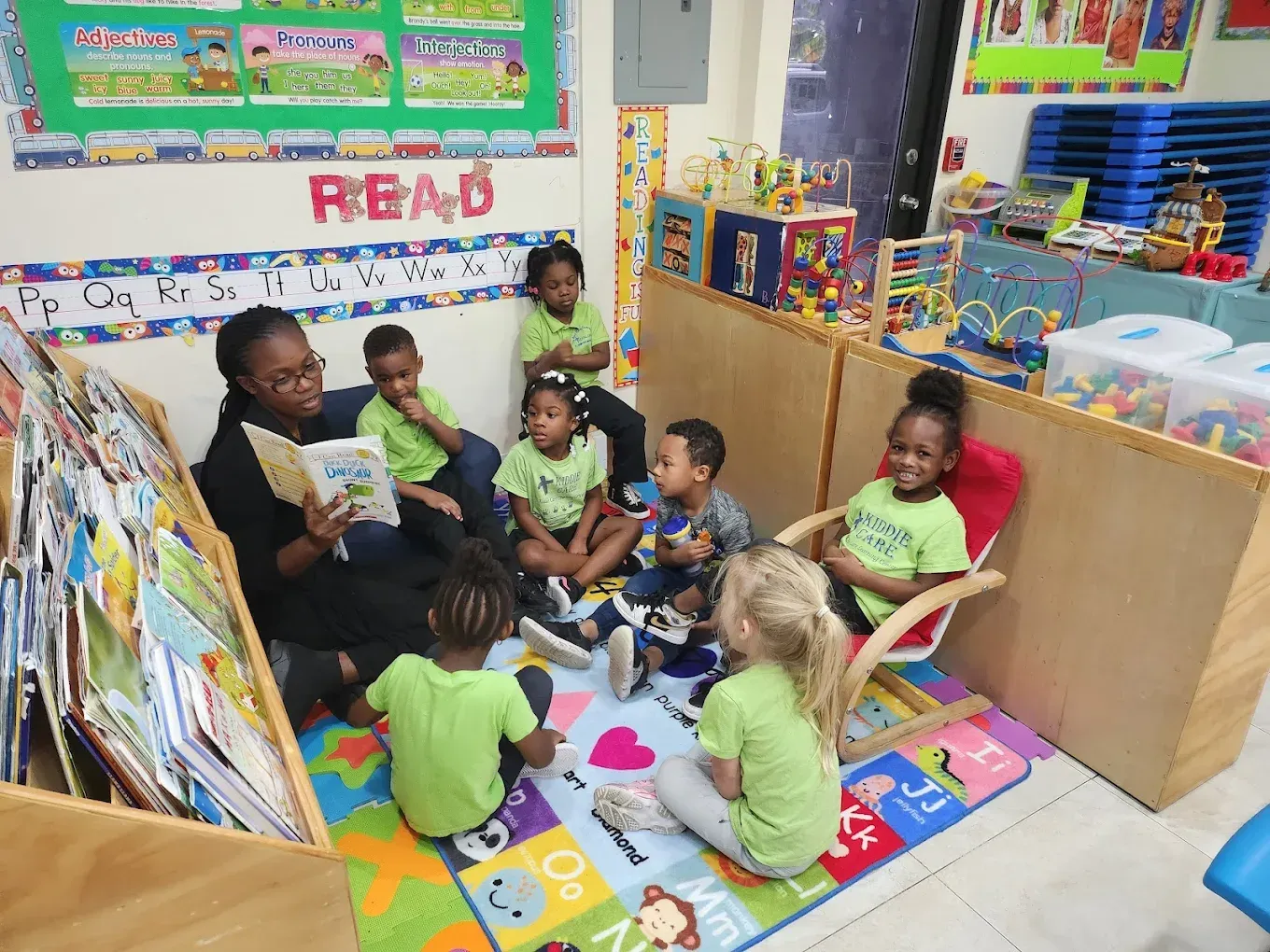 A teacher reads a book to a group of children sitting on a colorful alphabet rug in a preschool classroom.