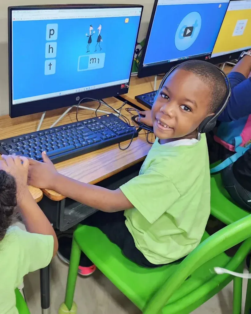 A student wearing headphones sits at a computer, smiling while completing a phonics learning activity on the screen.