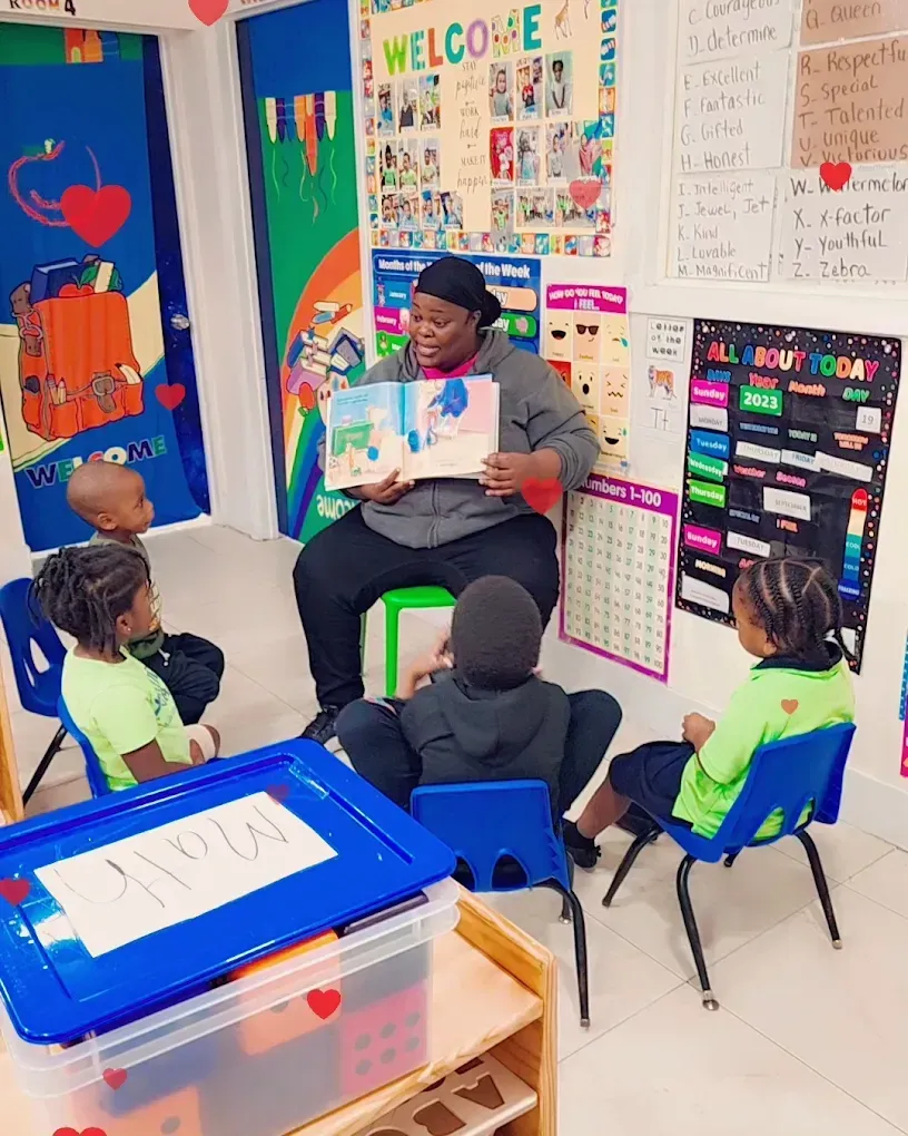 A person sits in a classroom reading a book aloud to four children seated in chairs around them.