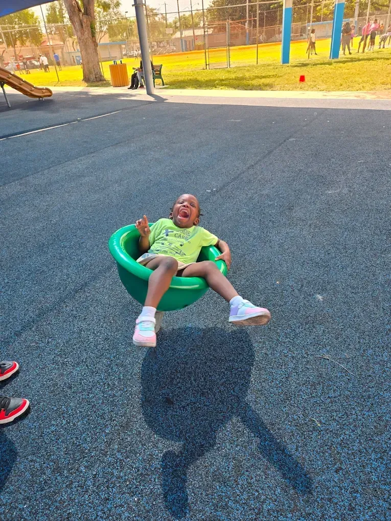 A child laughs while sitting in a green spinning playground seat on a paved surface.