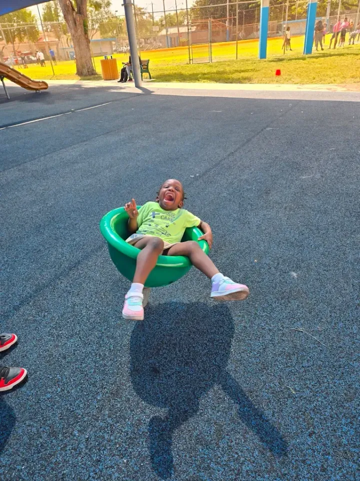 A child laughs while sitting in a green spinning playground seat on a paved surface.