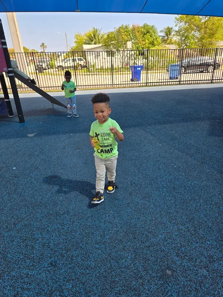 A child in a bright green shirt walks across a blue playground surface; another child stands in the background.