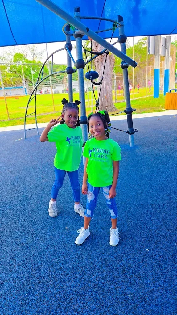 Two children in matching neon green t-shirts and patterned leggings pose on a blue playground surface under a canopy.