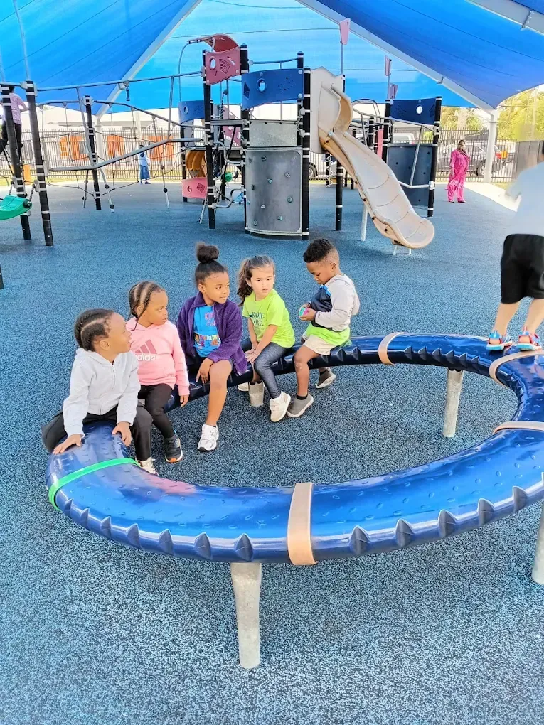 Four children sit on a curved blue playground structure under a large sunshade, with climbing equipment in the background.