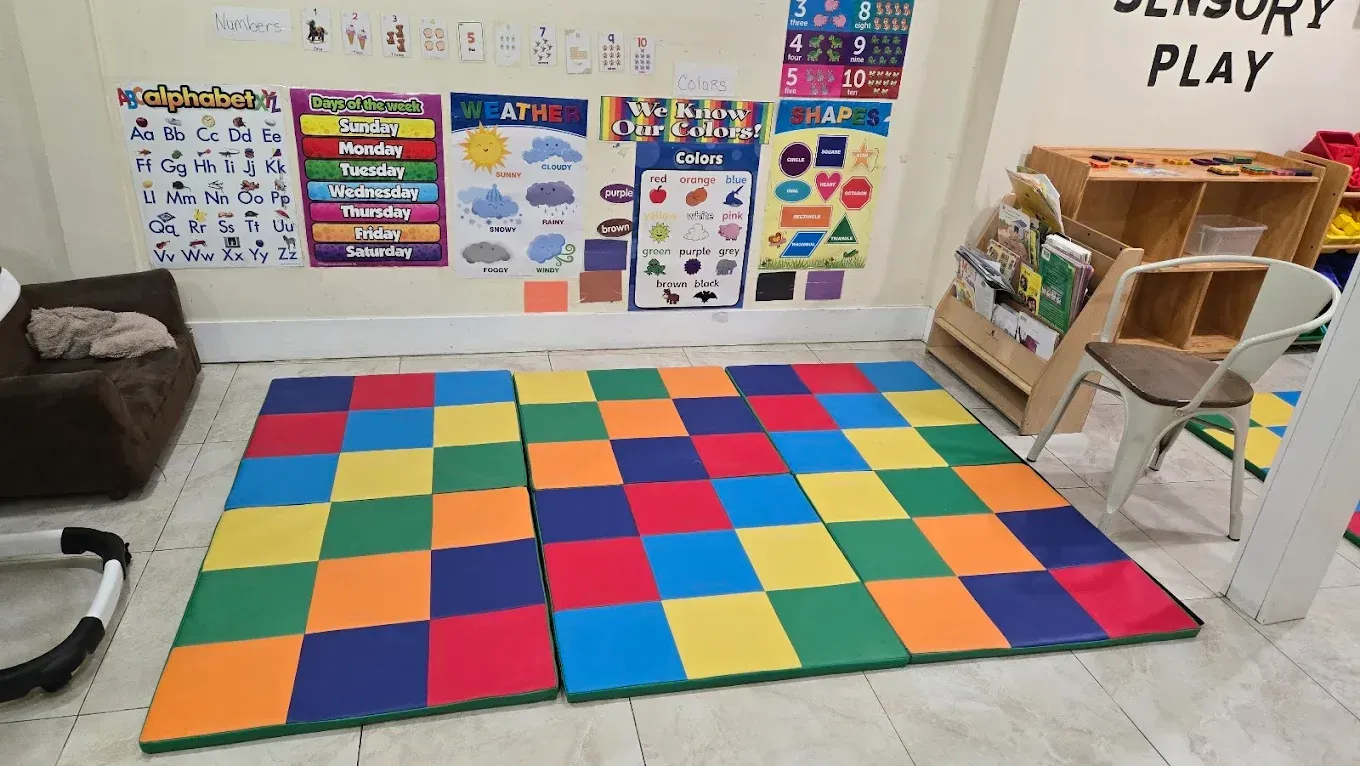 A colorful grid play mat on the floor of a classroom with posters and shelves in the background.