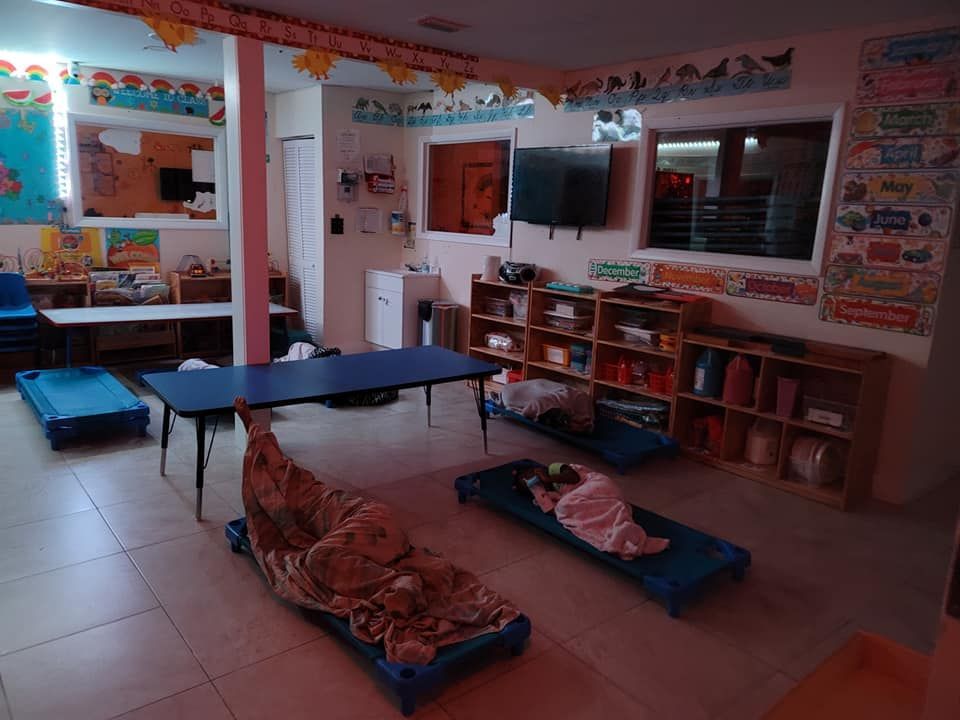 A preschool classroom with three sleeping mats on the floor, a central blue table, and wooden storage shelves.