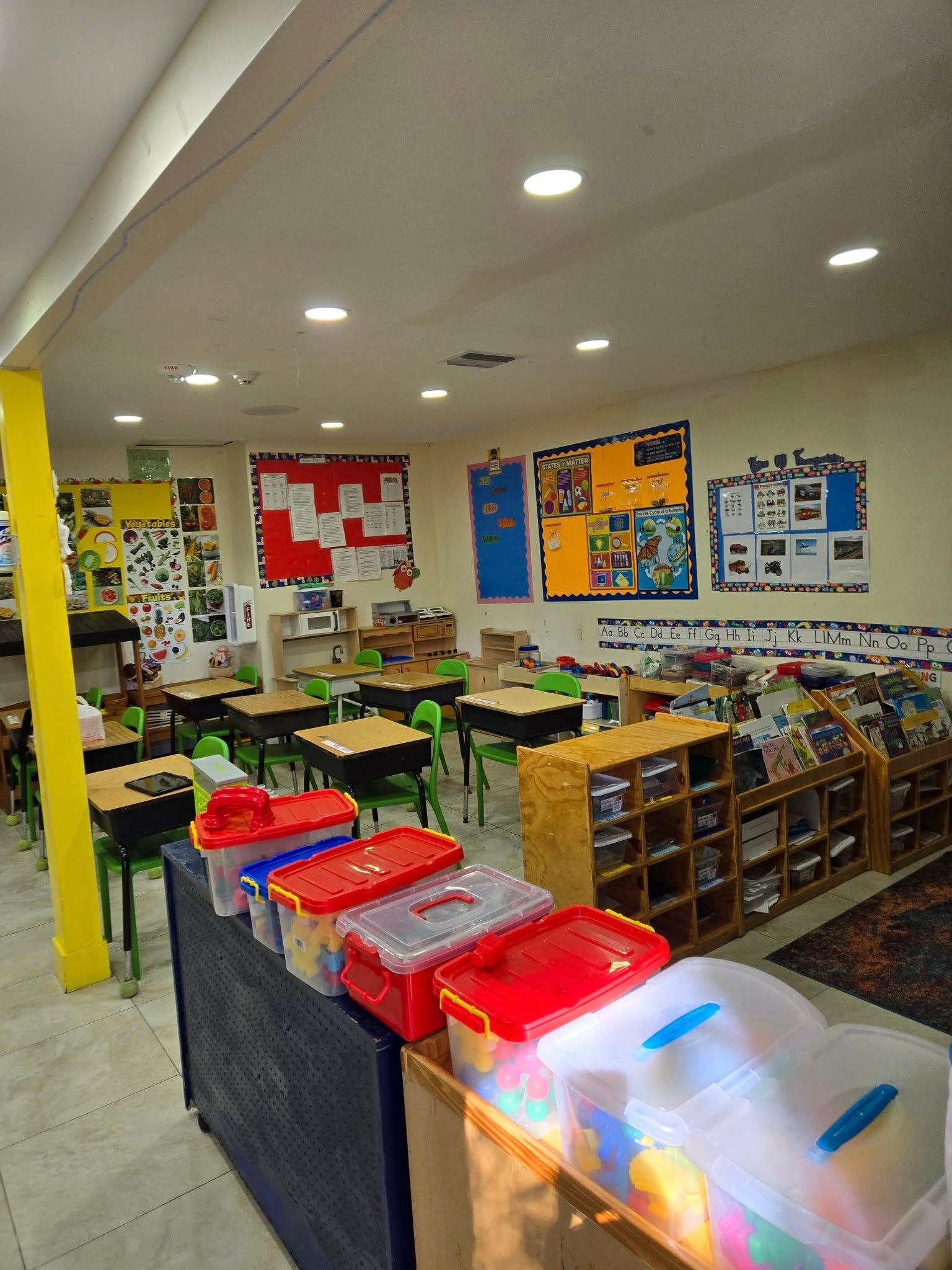 A classroom with rows of small student desks, learning posters on the walls, and colorful storage bins in the foreground.