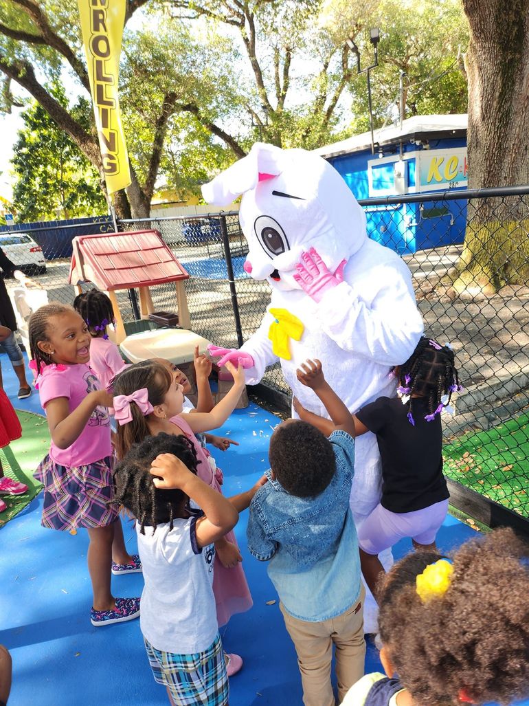 Children surround a person in a white Easter bunny costume on a blue outdoor playground surface.