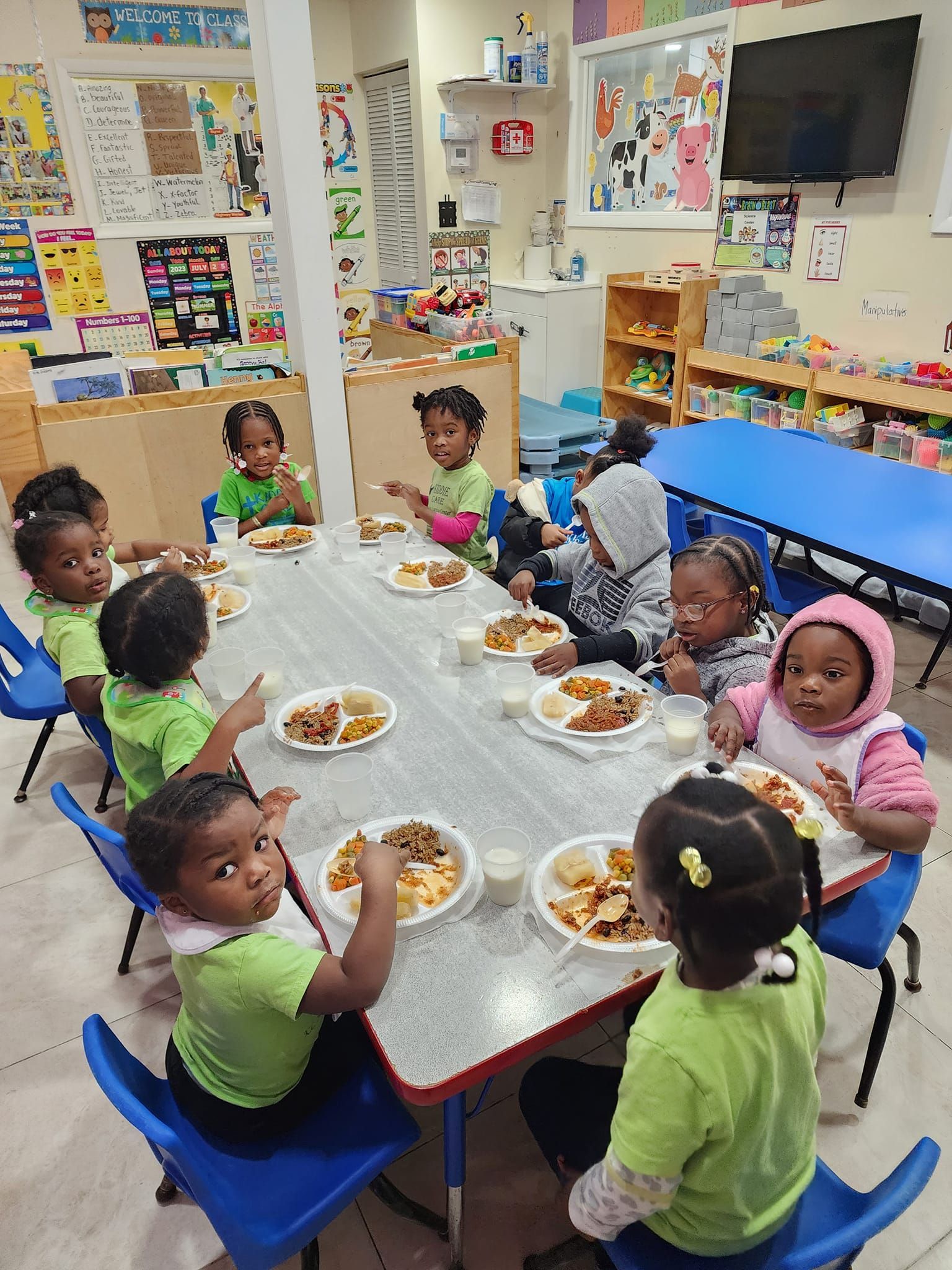 Children in green shirts sit at a table in a bright classroom, eating lunch from white plates with blue chairs.