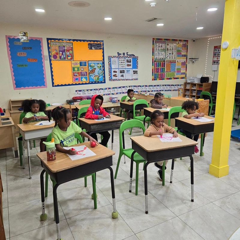 Children sit at individual desks in a brightly lit classroom, working on papers with crayons and markers.