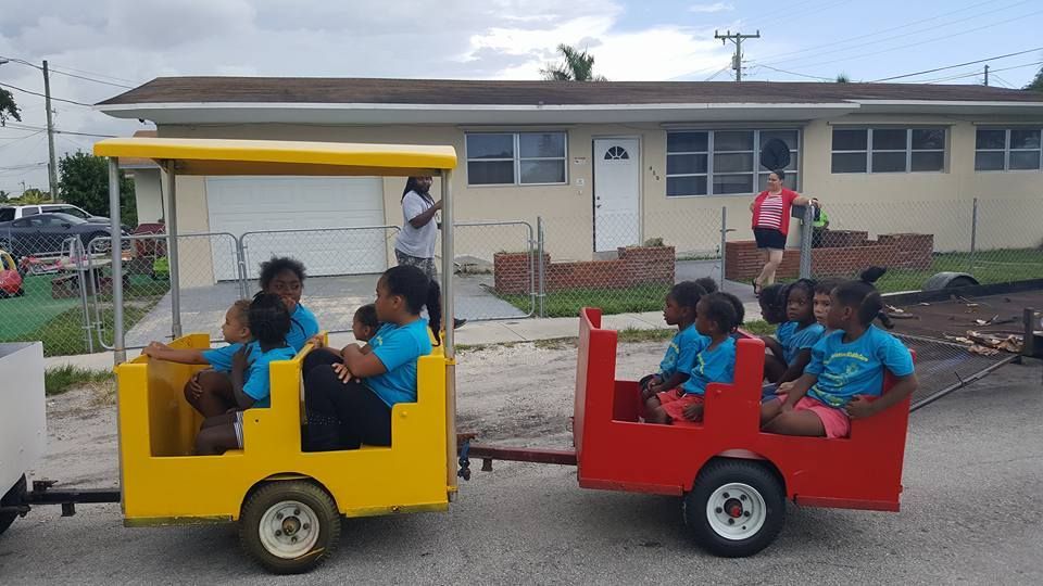 Children ride in a two-car miniature train painted yellow and red, parked on a street in front of a house.