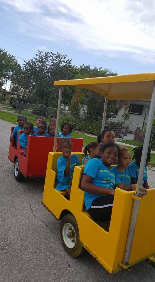 A group of children in matching blue shirts riding in two connected, colorful open-air wagons outdoors.