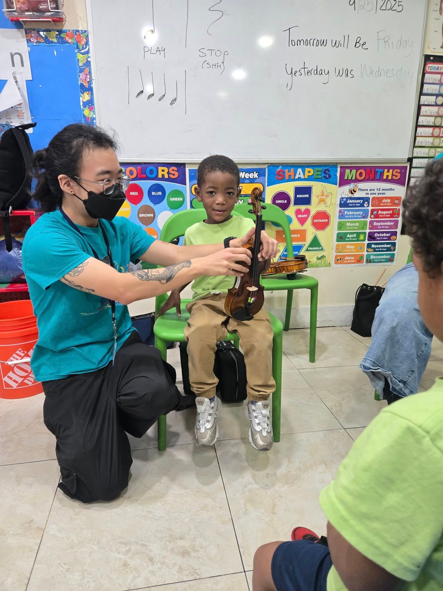 A person in a turquoise shirt helps a child in a green shirt hold a violin in a classroom with educational wall posters.