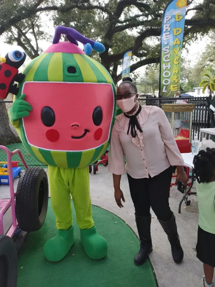 A person in a light pink blouse and dark pants poses with a Cocomelon mascot costume outdoors at a preschool event.
