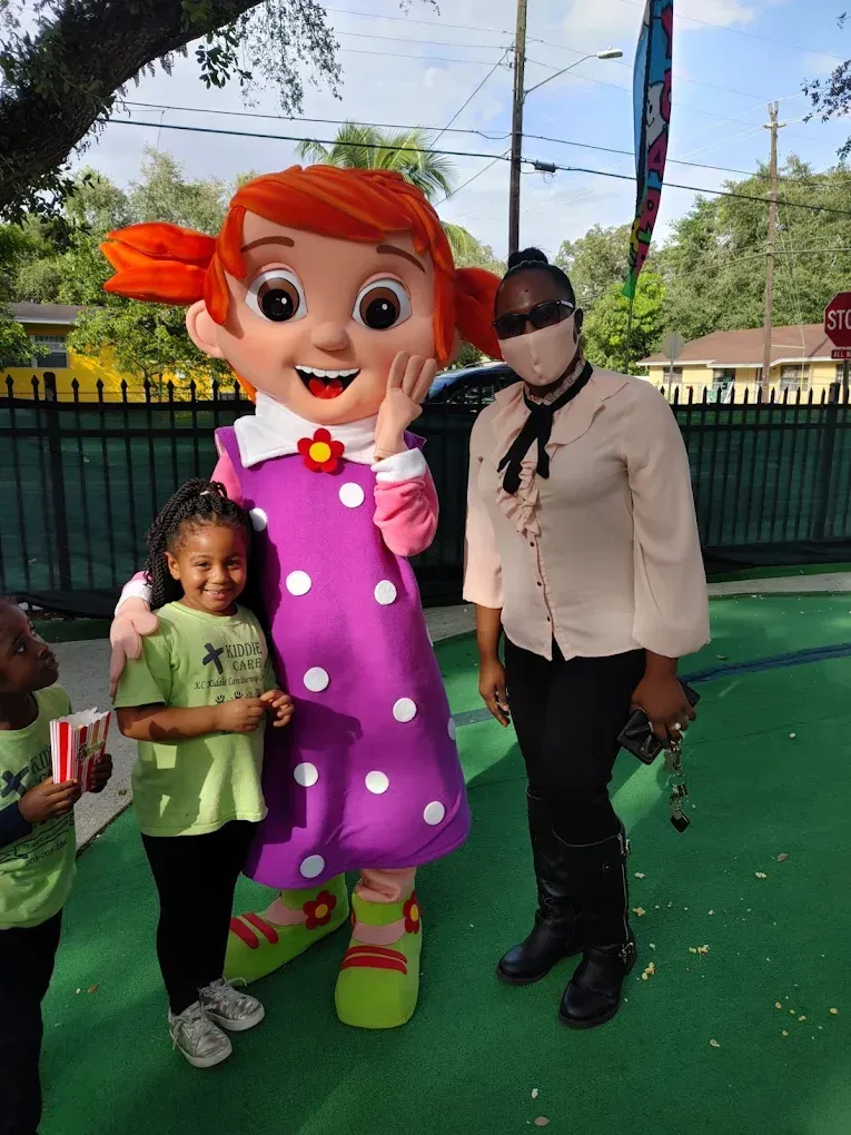 A child, a mascot in a purple dress, and an adult wearing a face mask pose together outdoors on a green surface.
